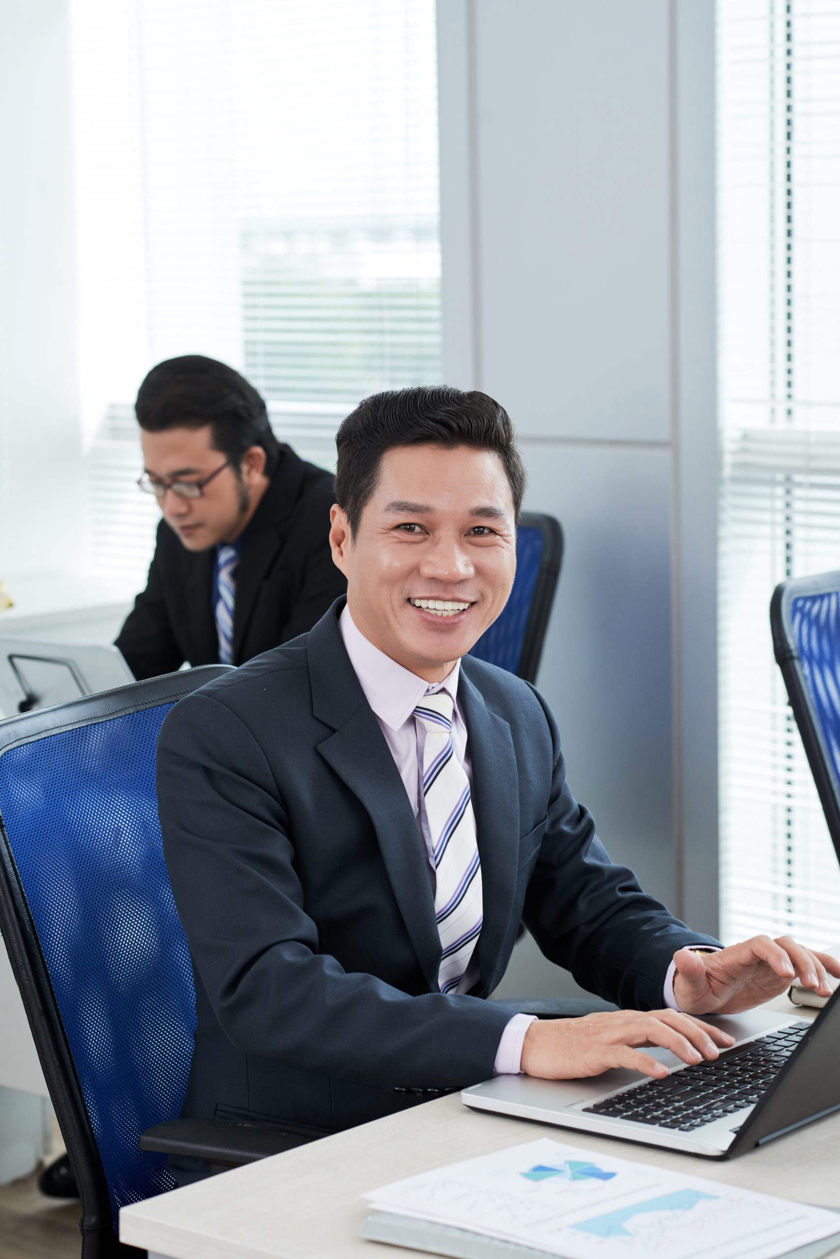 Waist-up portrait of middle-aged Asian manager wearing suit posing for photography with smile while working on promising project with modern laptop, interior of open plan office on background